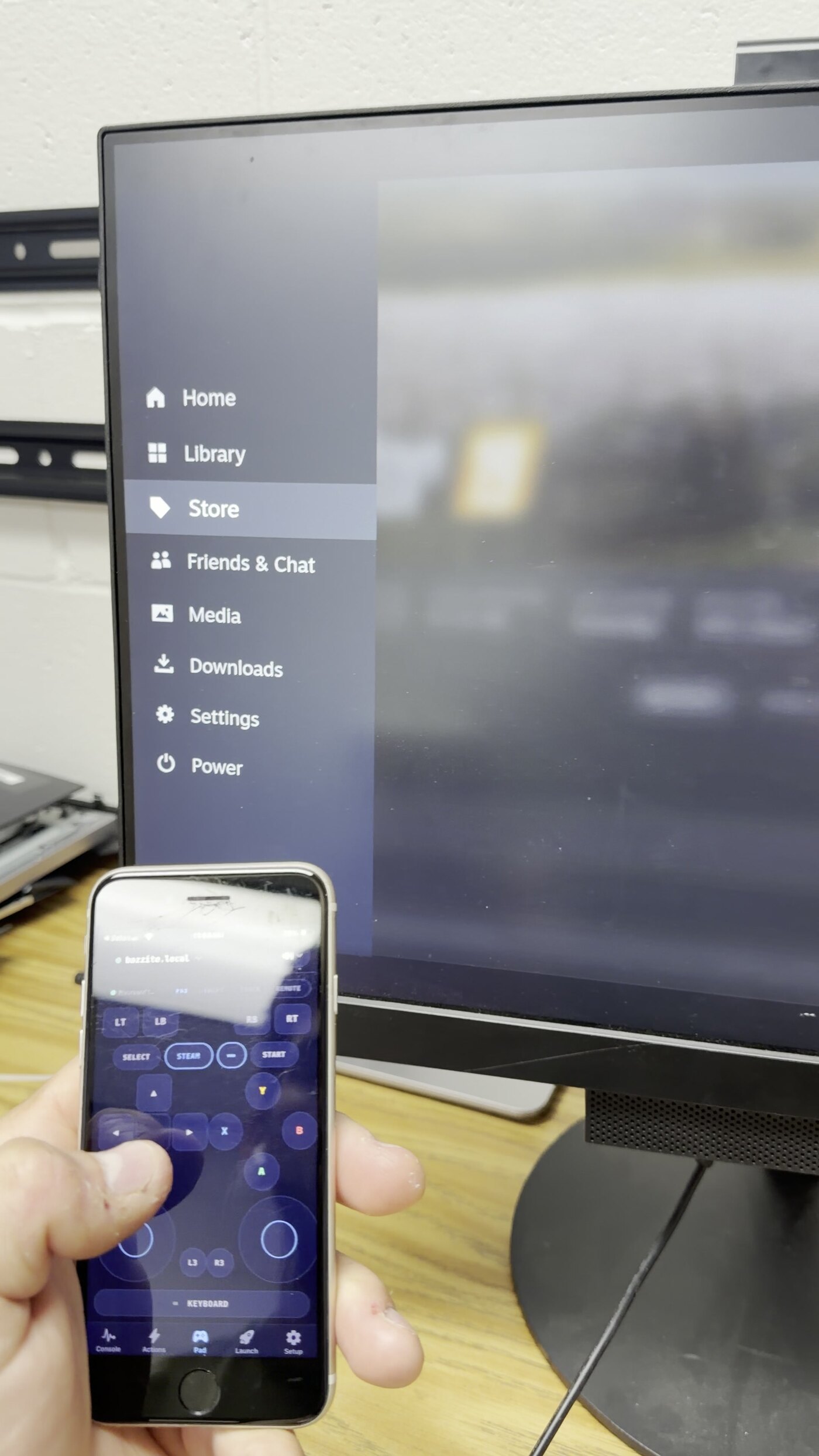 A phone showing the Couchside gamepad while the TV behind it shows the Steam menu the phone is driving